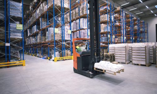 Worker operating forklift machine and relocating goods in large warehouse center.